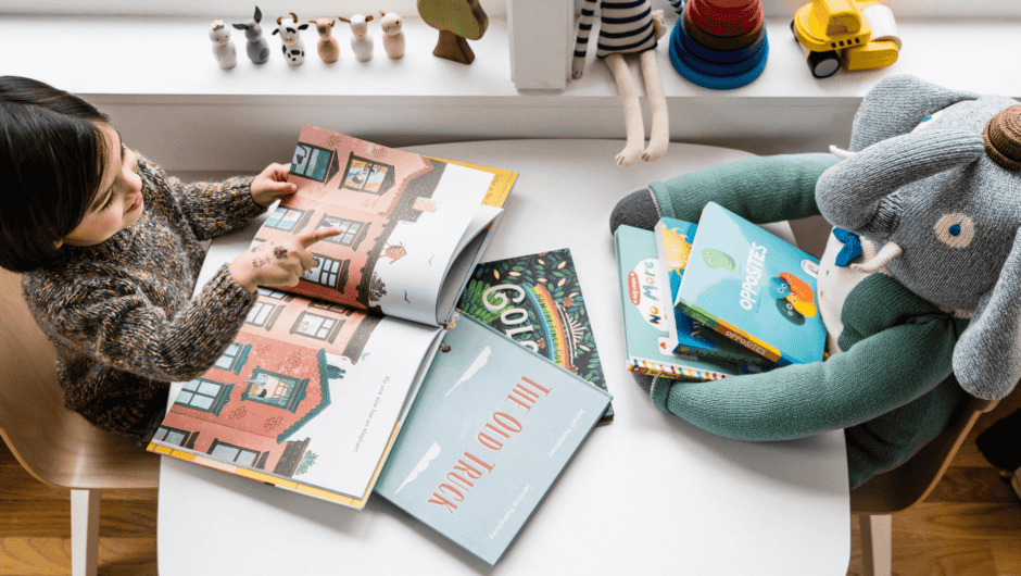A little boy reads picture books at a tiny table with his elephant plush toy.