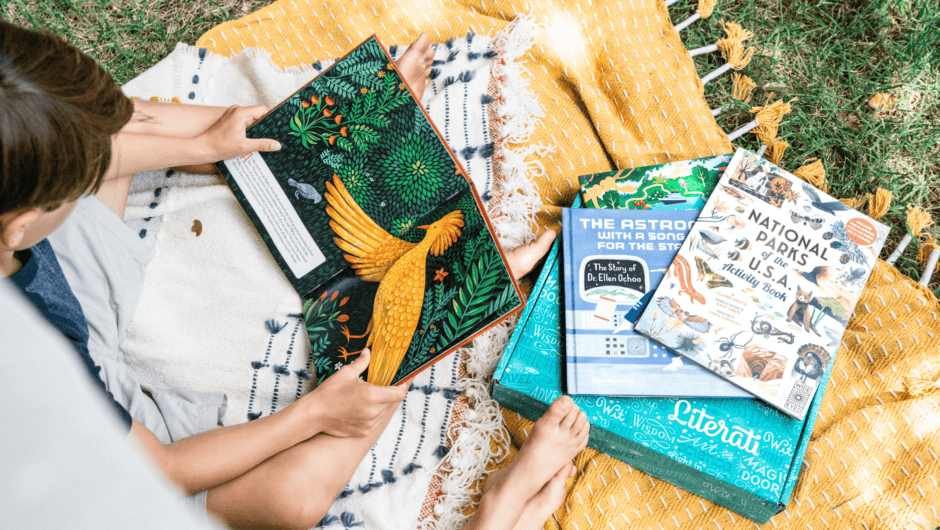 A boy sits on a yellow blanket with a colorful picture book open, and two more books on a Literati box.