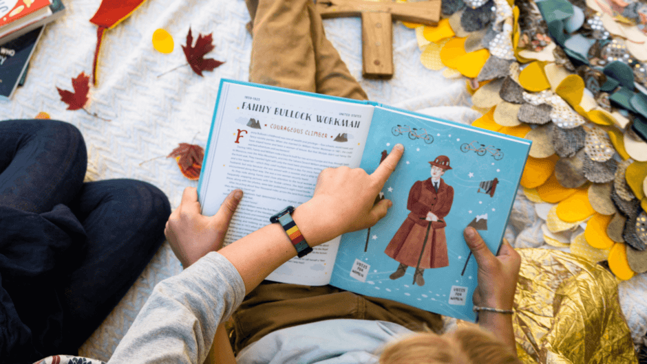 A child points to something on the page of an open book, while sitting on a blanket with fall leaves.