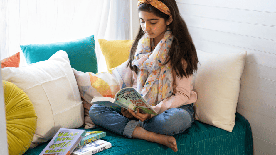 A girl sits crossed legged in a picture window reading a book.