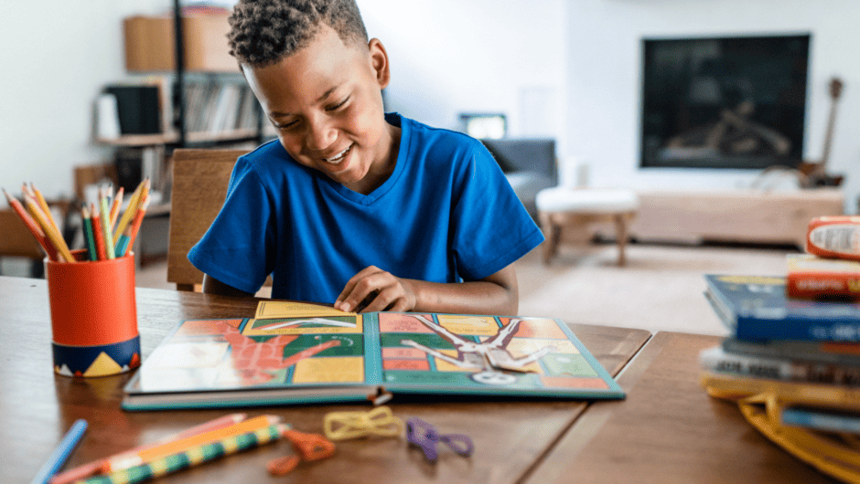 A young boy is smiling at the kitchen table while reading a book